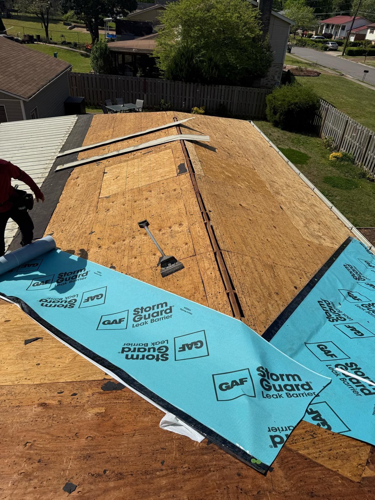 Roofer installing blue roofing underlayment on an exposed wood roof with adjacent yard and houses.