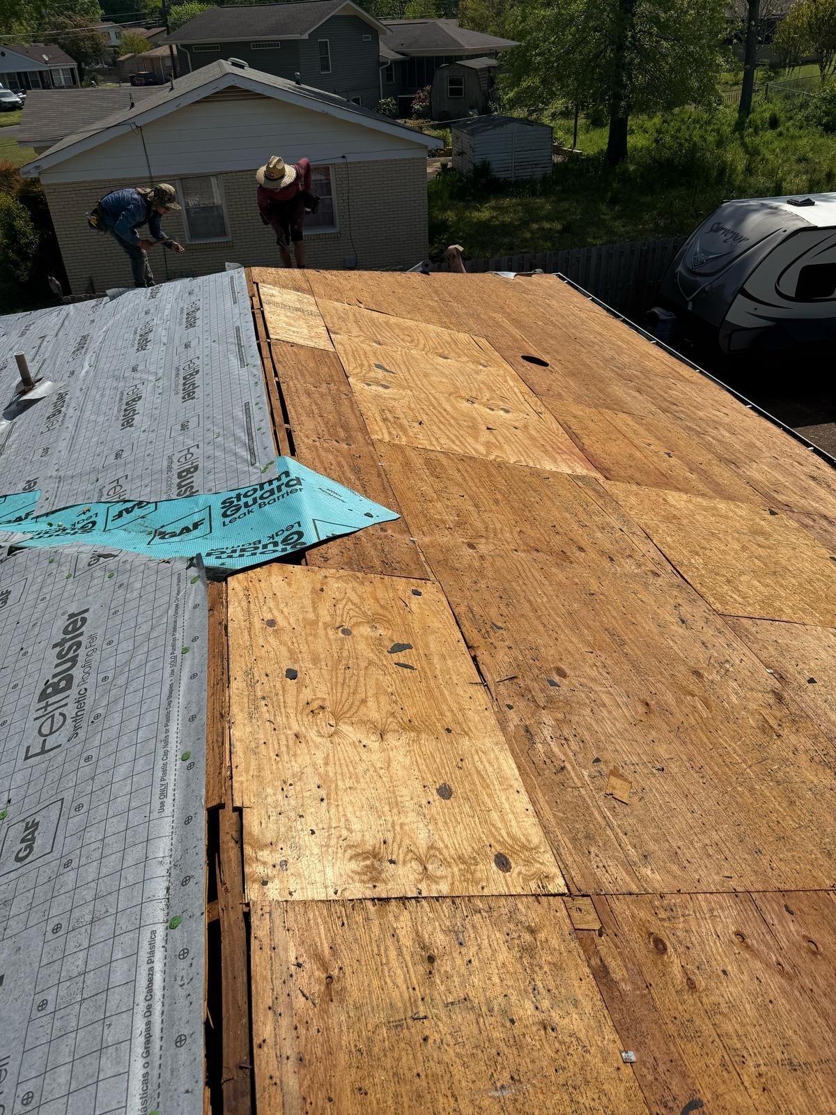 Roof partially covered with plywood, and blue underlayment; workers on roof in the background.