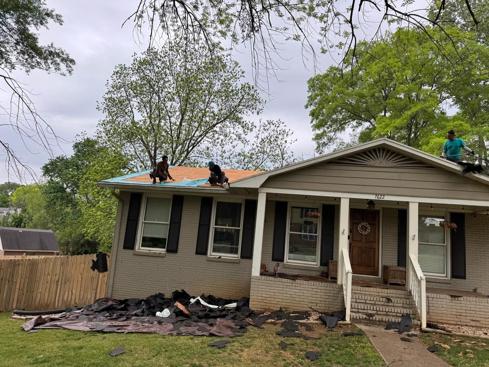 Roofers working on a house with removed shingles. Debris on the ground, trees in the background. Cloudy sky.