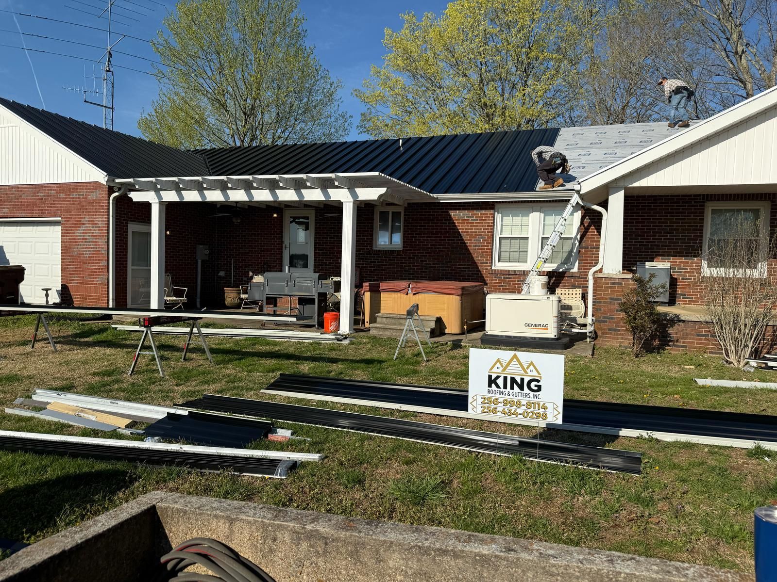 Workers installing a black metal roof on a brick house. Tools and materials are in the yard.