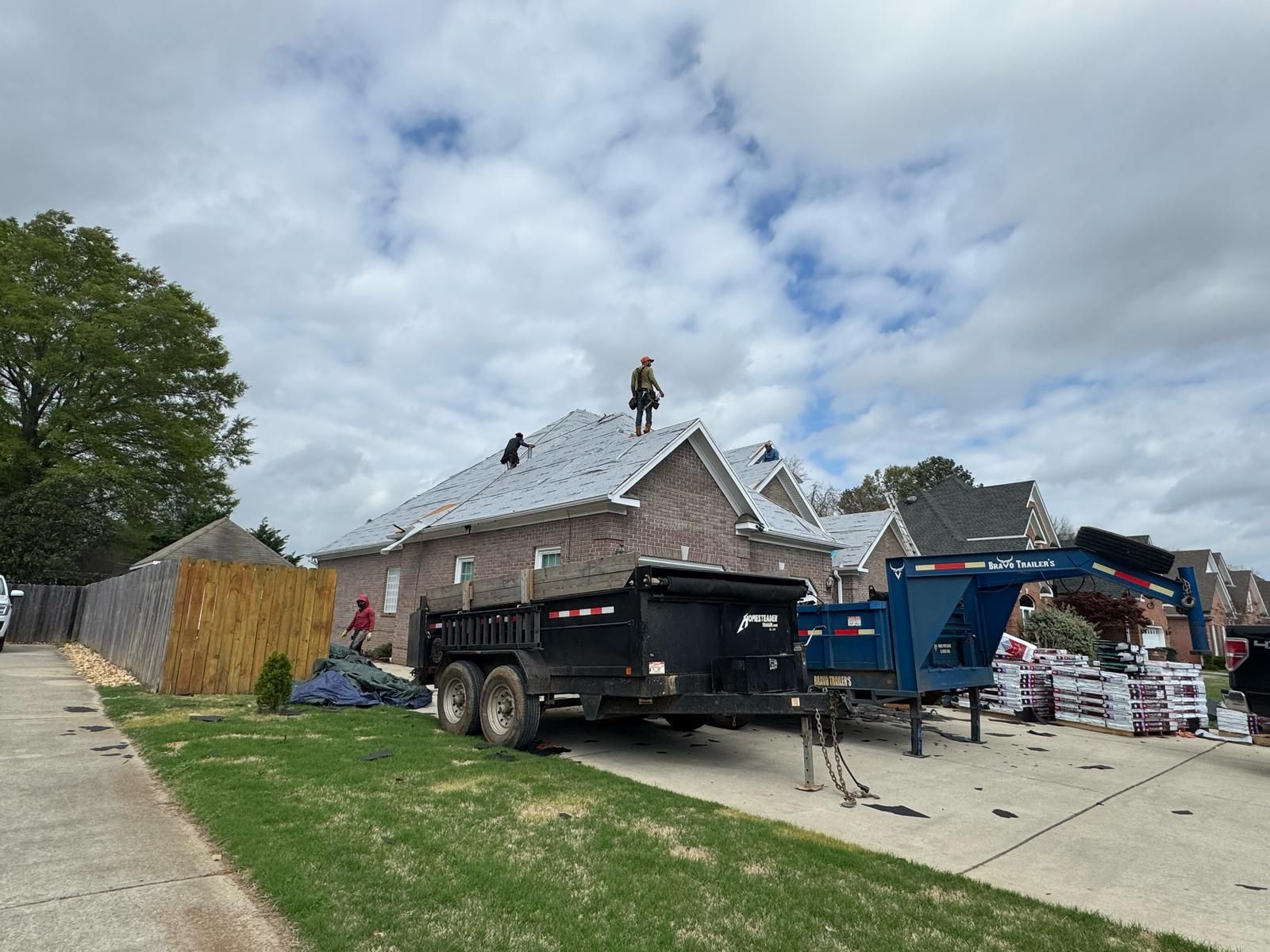 Roofing crew working on a house with a black trailer and supplies parked in front.
