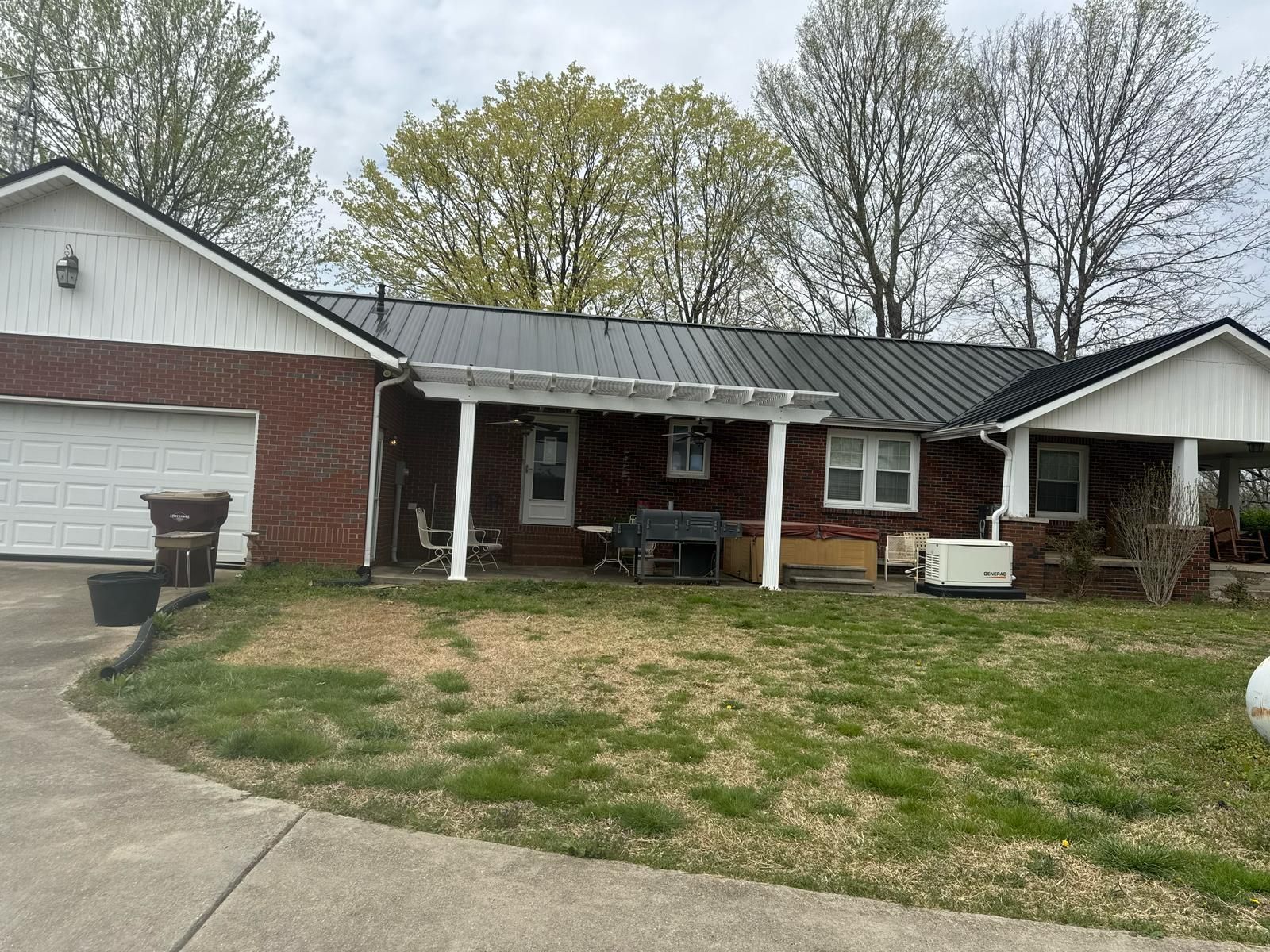 House with brick garage, black metal roof, porch, and a lawn. Trees in the background, overcast sky.
