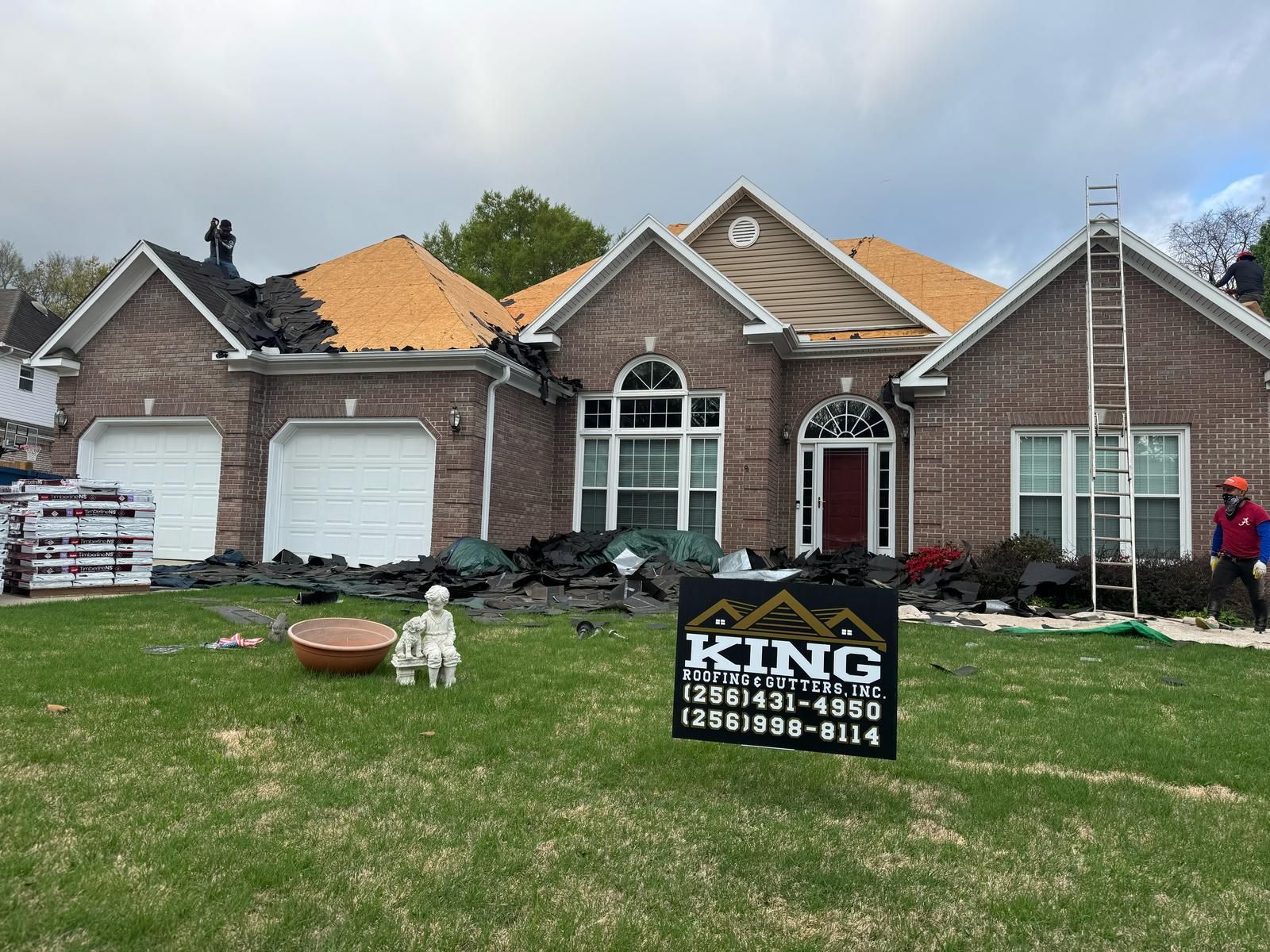 House roof being replaced, shingles removed. Ladder, King Roofing sign on lawn. Brick exterior, overcast sky.