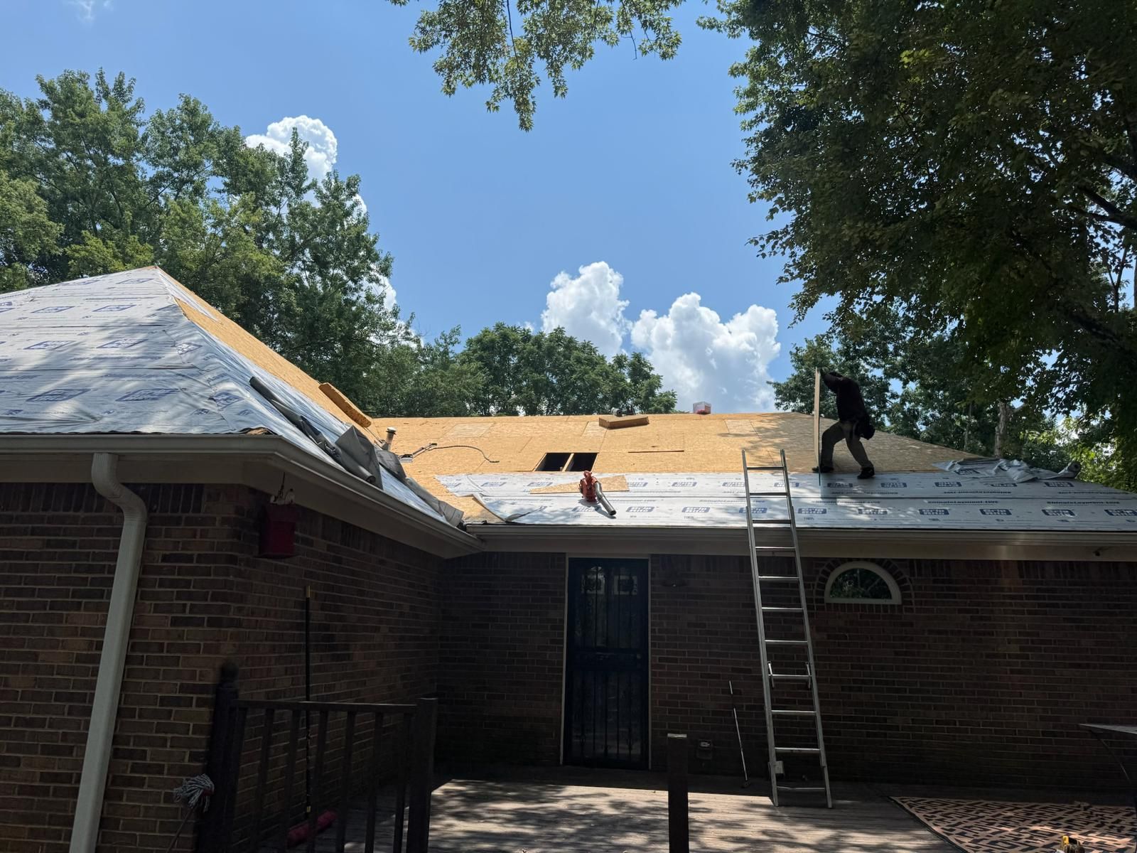Roofers working on a house, replacing shingles. Ladders, blue sky, trees.