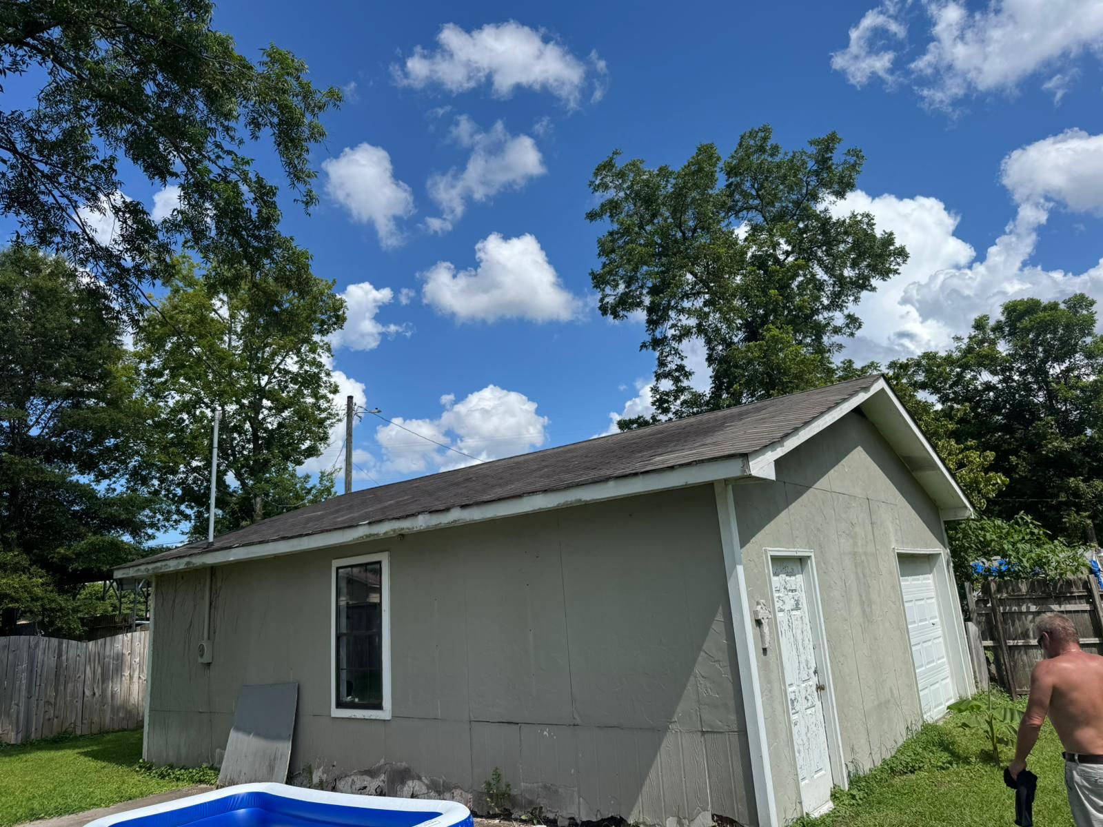 Gray building with dark roof under a partly cloudy blue sky, trees in background, small pool in foreground.