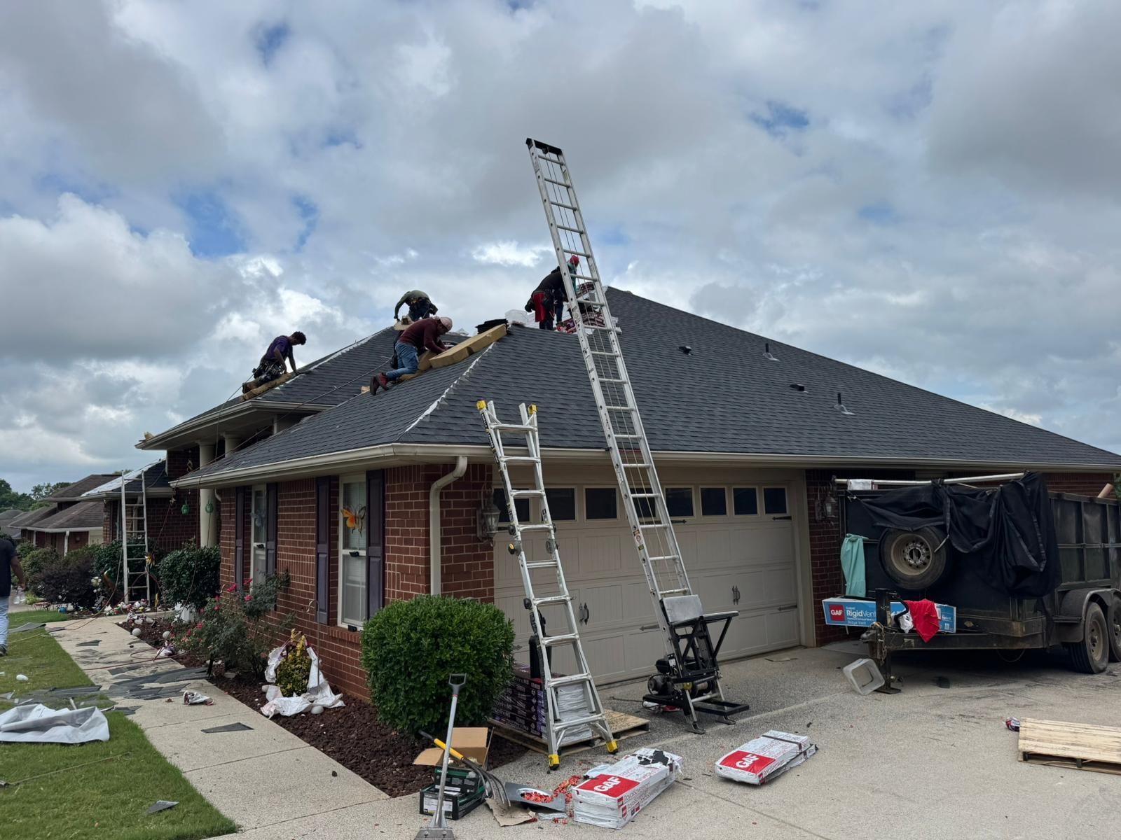 Roofers working on a house with a dark roof; ladders and construction supplies are visible. Overcast sky.