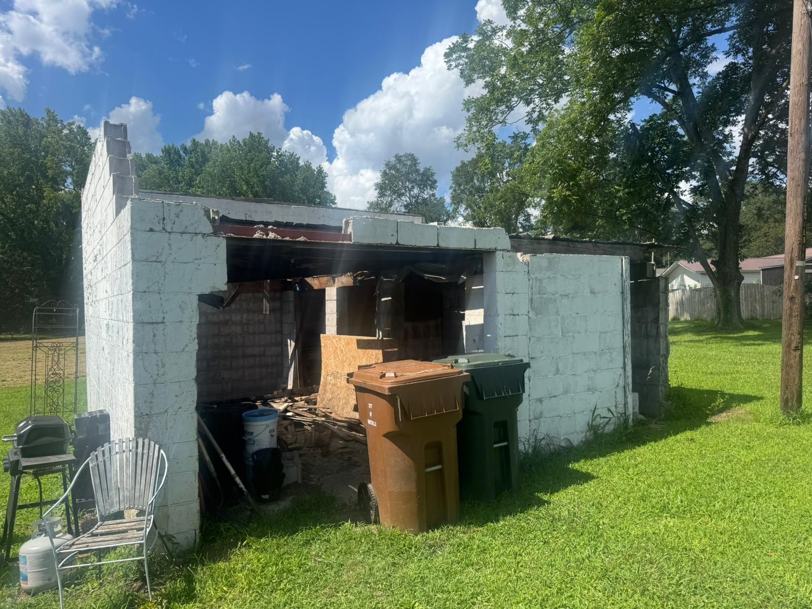 Dilapidated white brick structure with open front, two trash cans, and overgrown lawn.