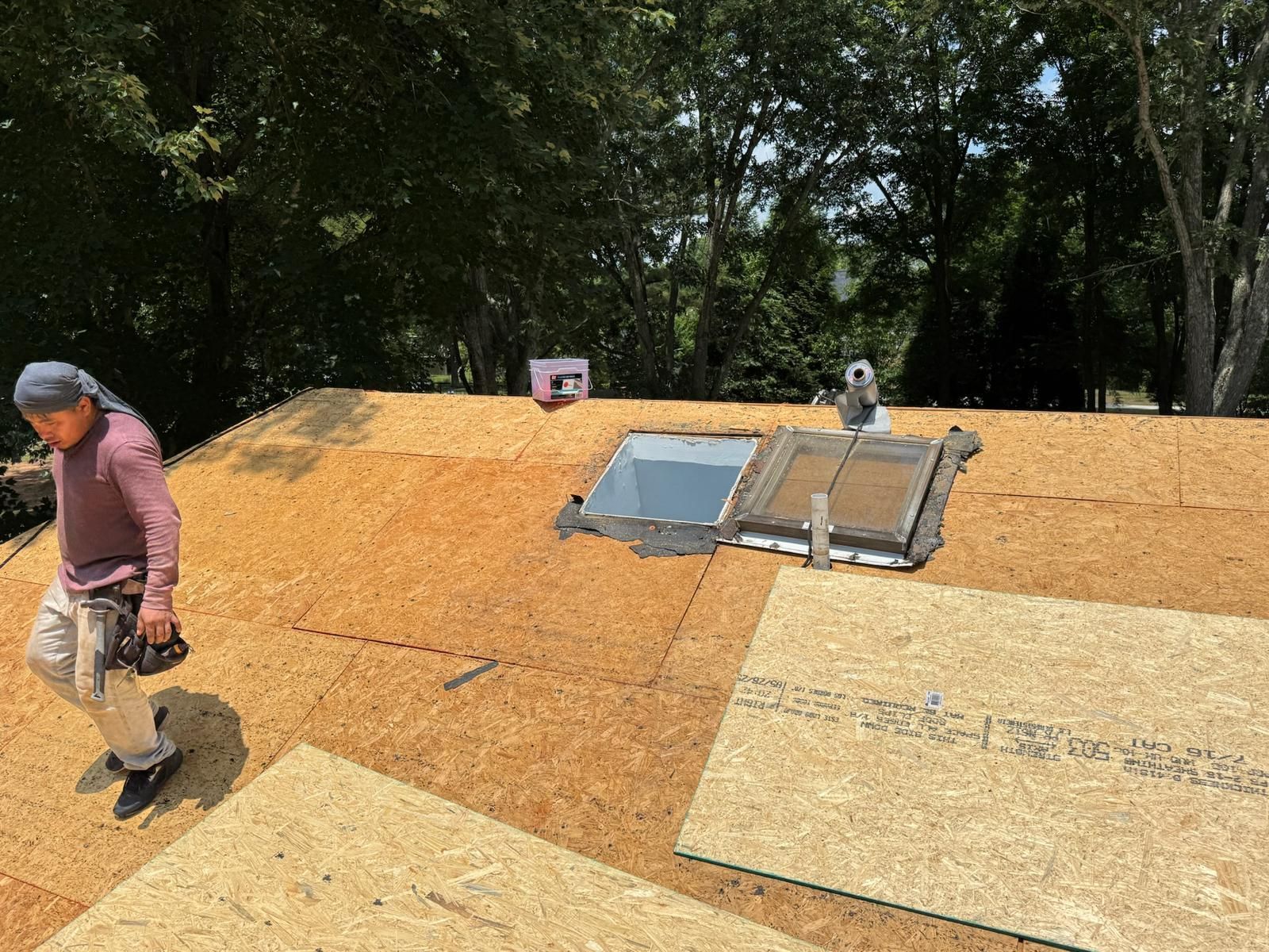 Roofer walking on a roof with plywood, preparing for new shingles. Sunny day, trees in background.