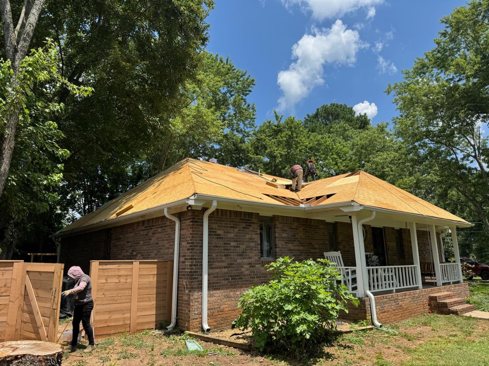 Workers removing roof shingles from a one-story brick house on a sunny day. A person works near a wooden fence.