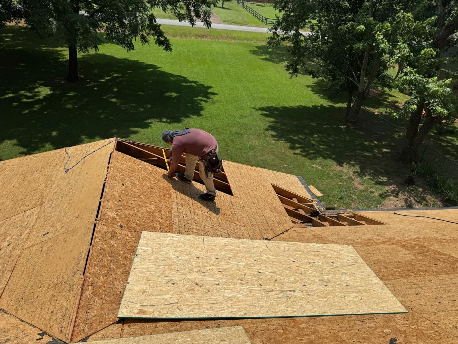 Roofer on a rooftop, installing wooden boards. Green grass and trees visible in the background. Sunny day.