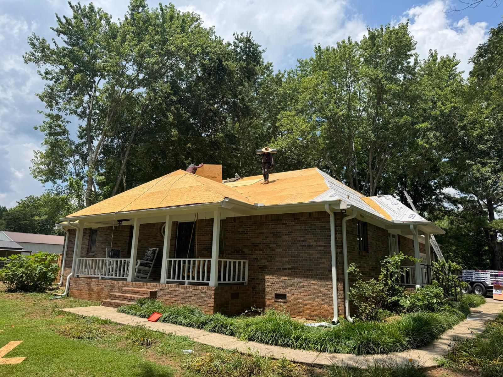 House undergoing roof replacement; exposed plywood, brick exterior, porch with white columns, ladder, trees in background.