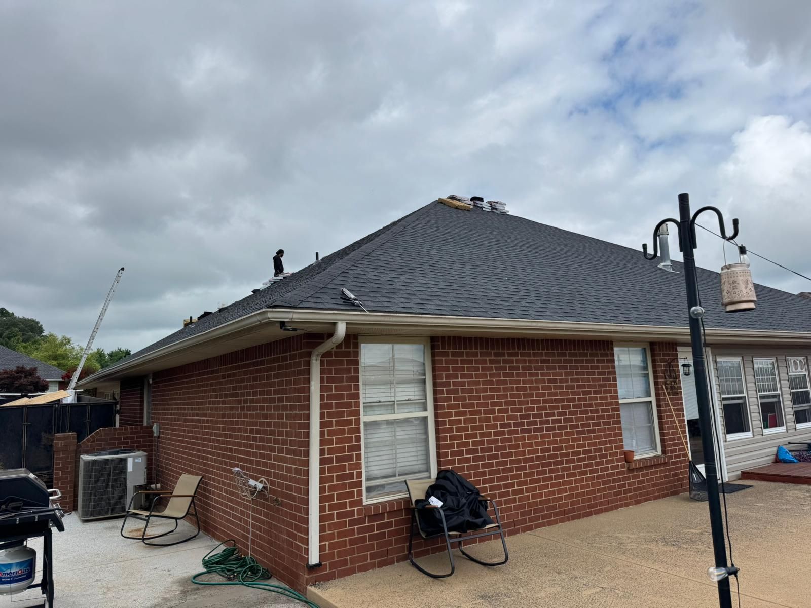 Brick house with dark roof under cloudy sky.  Gutter, windows, outdoor furniture, and a grill are visible.