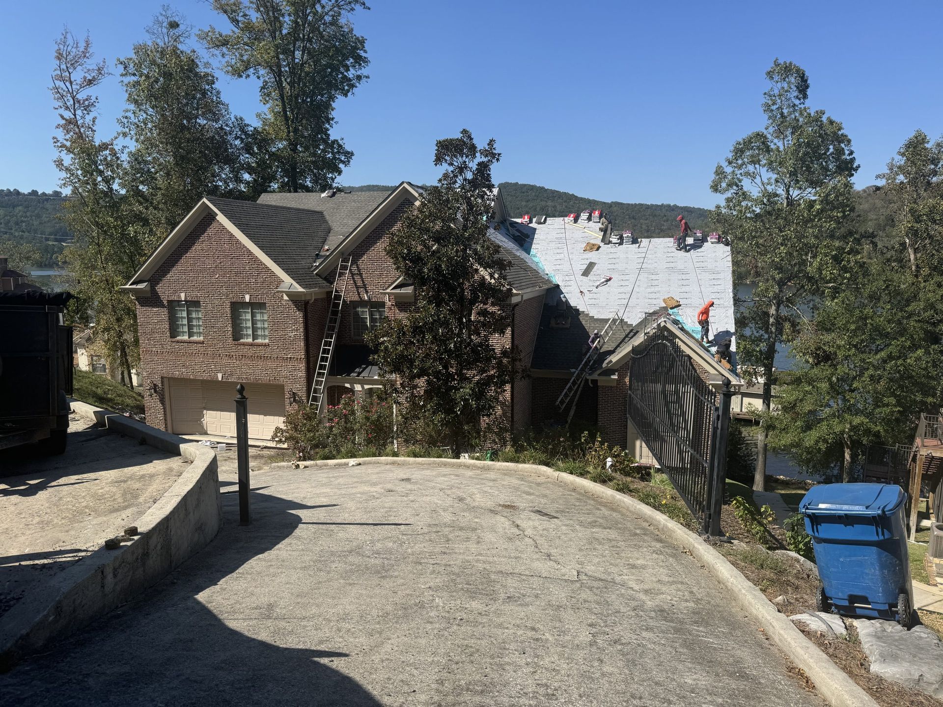 House with roof partially removed, construction workers on roof. Driveway, trees, and blue trash can in front.