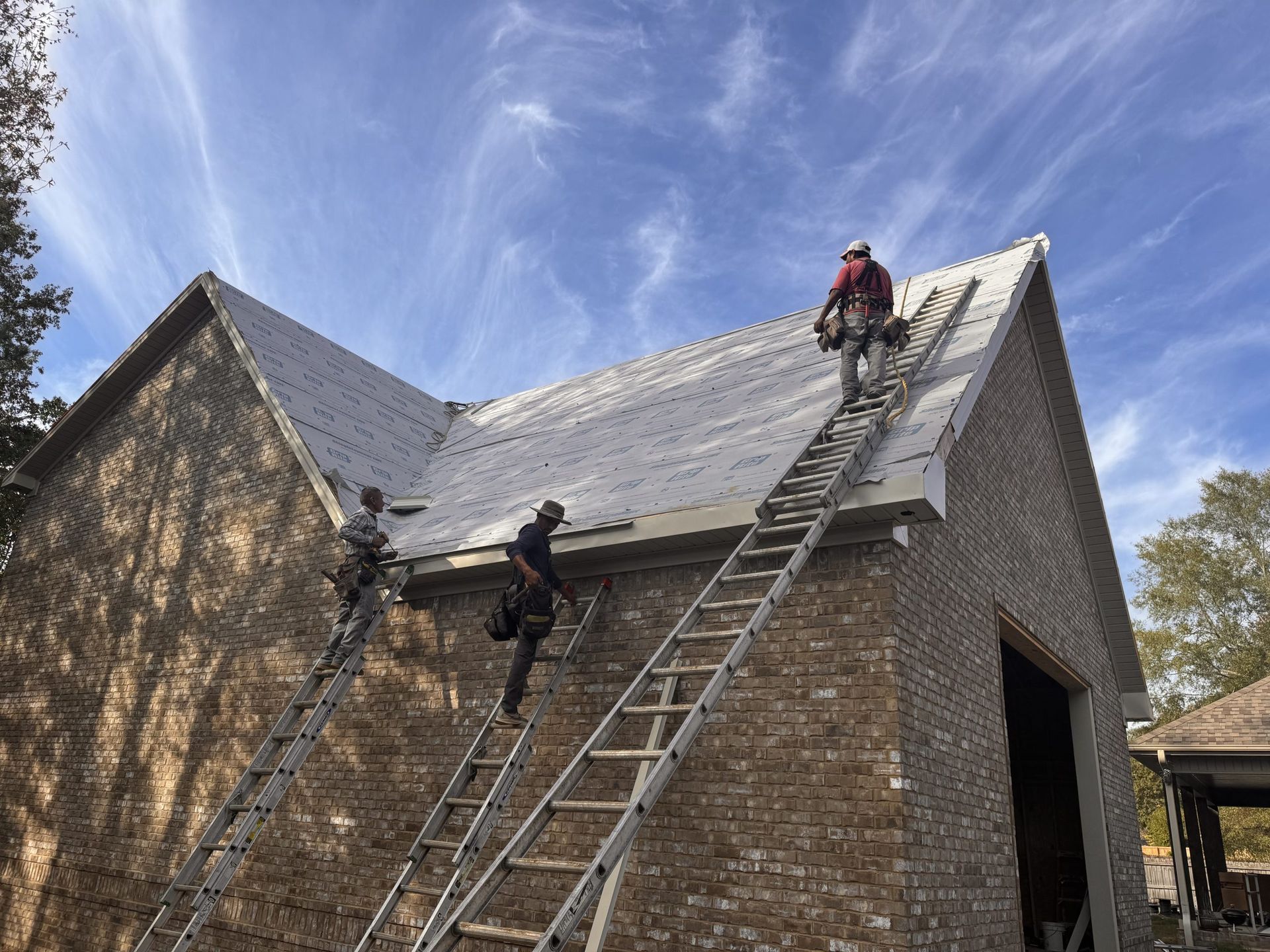 Three workers on ladders installing a roof on a brick building, blue sky backdrop.