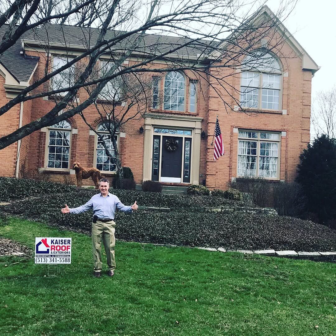 A man is standing in front of a large brick house holding a sign.
