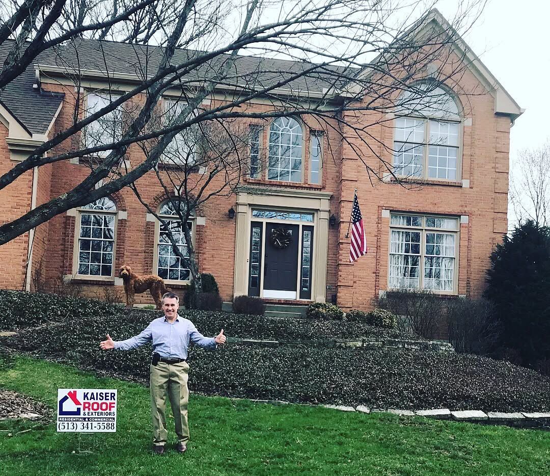 A man is standing in front of a large brick house holding a sign.