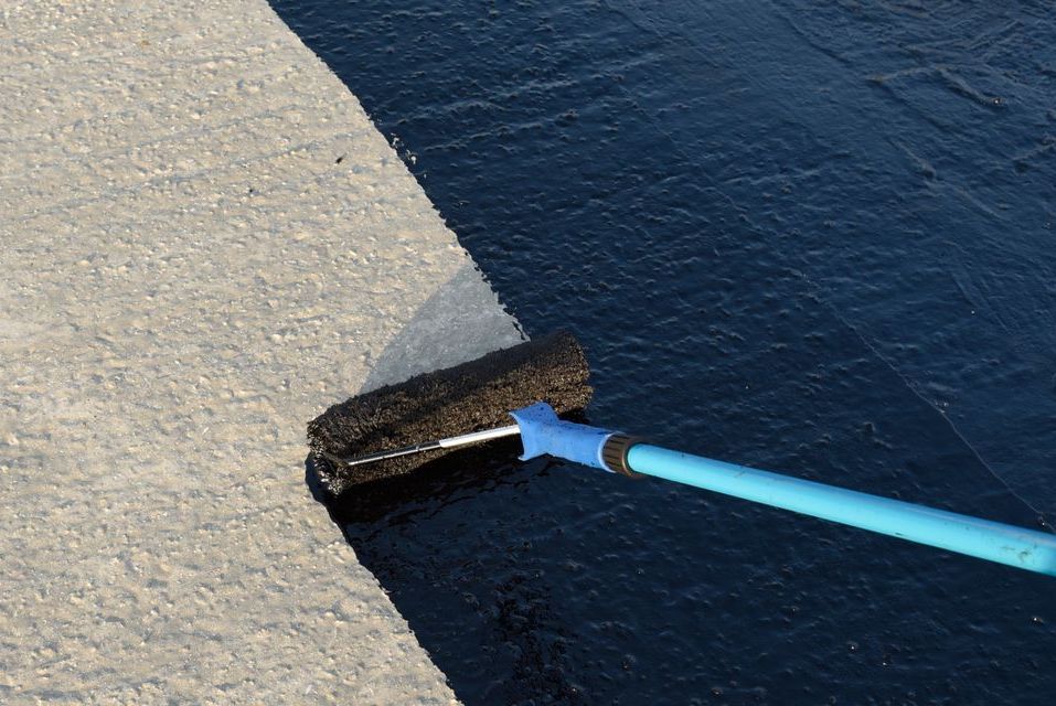A person is using a broom to spread black paint on a concrete surface.