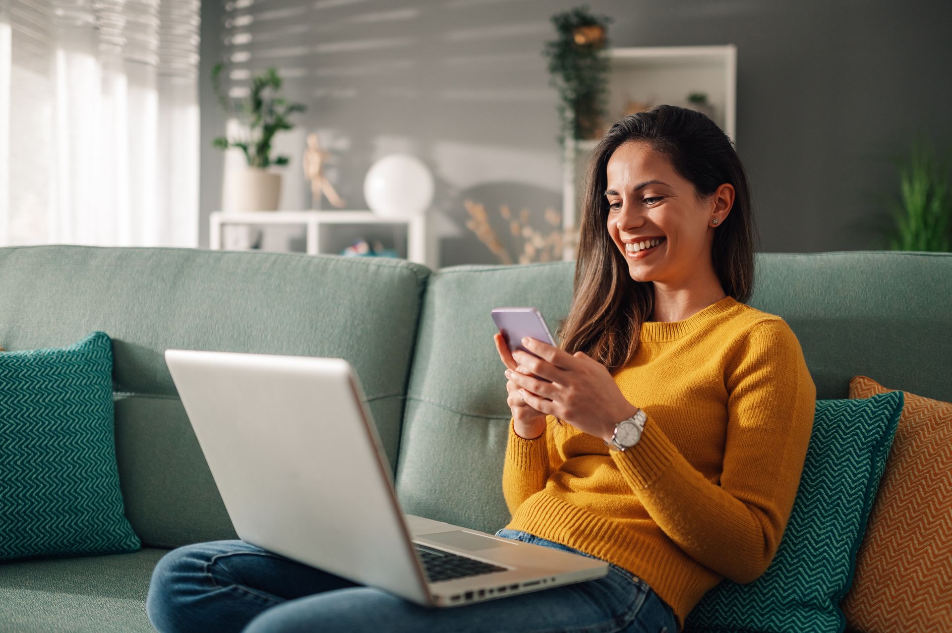 A woman is sitting on a couch using a laptop and a cell phone.