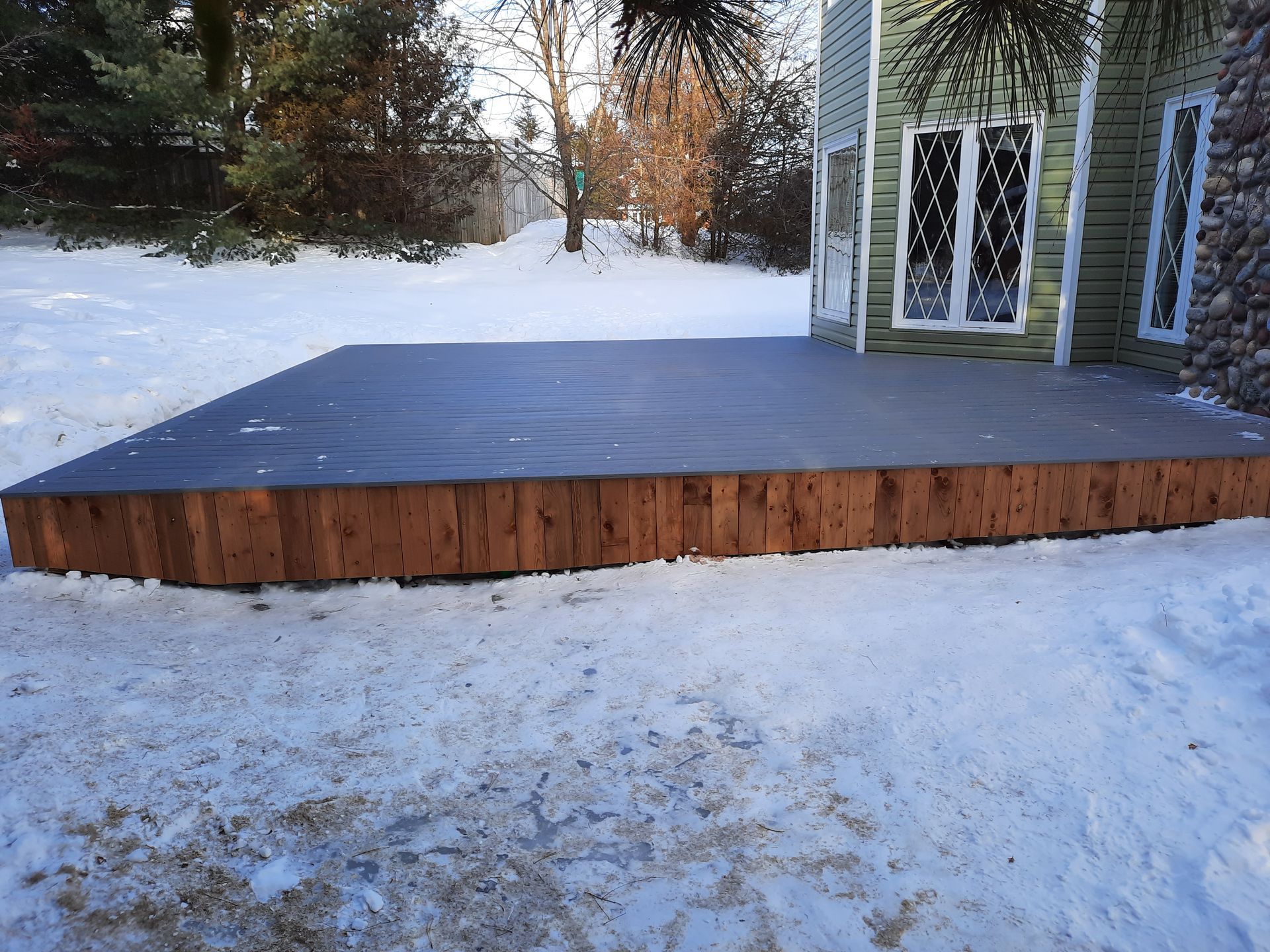 A large wooden deck is sitting in the snow in front of a house.