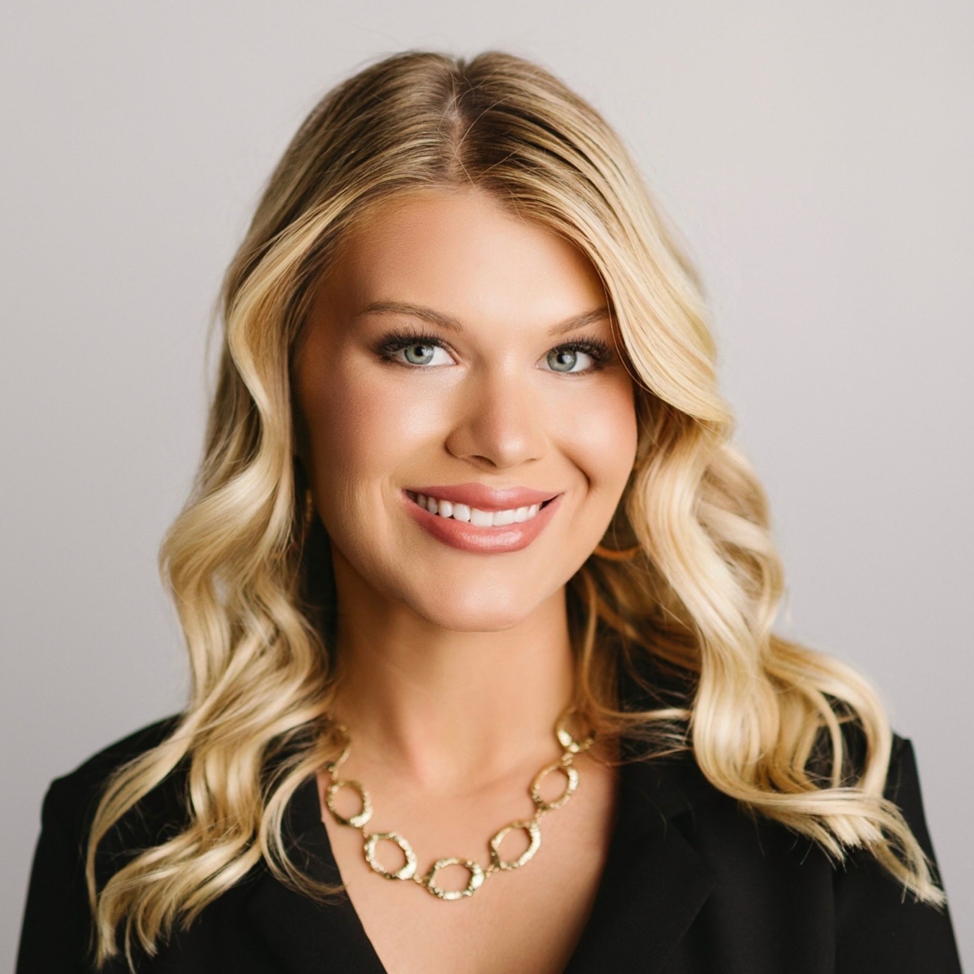 Blonde woman wearing a black blazer and gold necklace smiles. Studio portrait.
