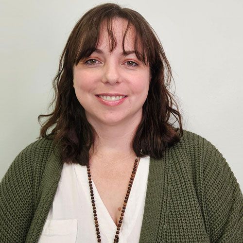 Woman with brown hair smiling, wearing a white shirt and olive green cardigan, beaded necklace.