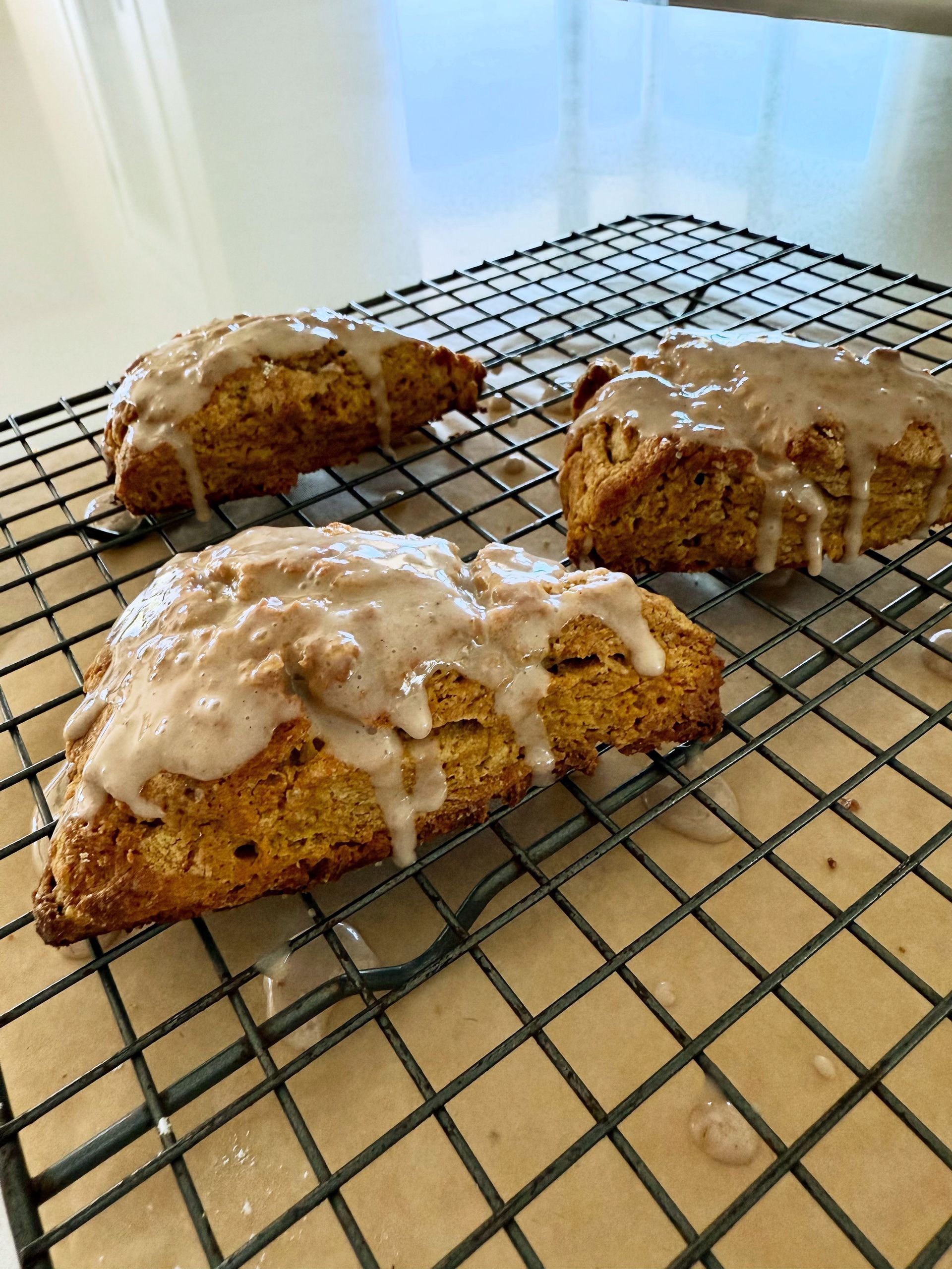 Three glazed scones on a wire rack, golden brown with dripping icing, on a light-colored countertop.