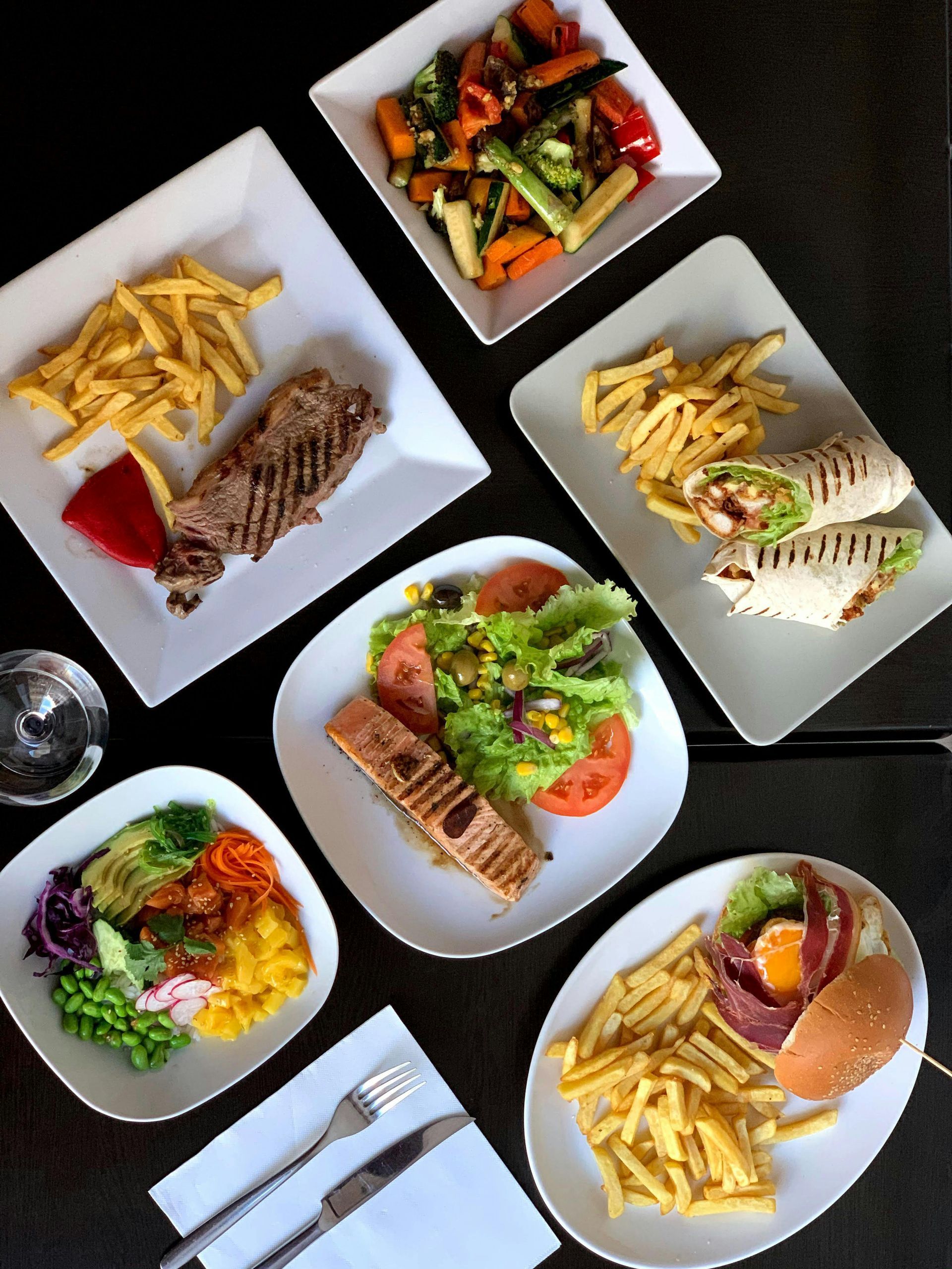 A top-down view of six varied dishes, including steak, salad, wraps, and a burger, arranged on a dark table.