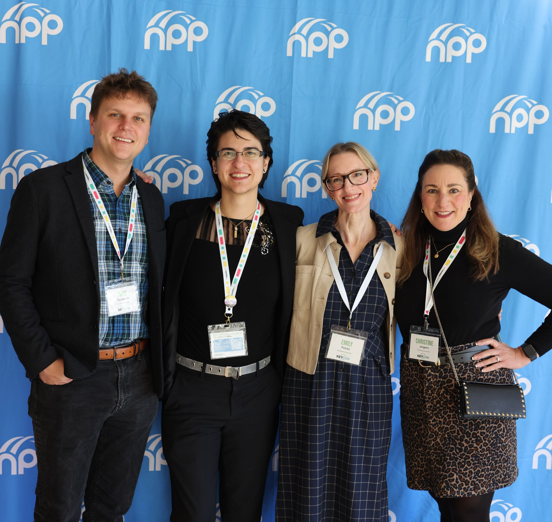 Four smiling people stand side-by-side in front of a blue banner with a recurring