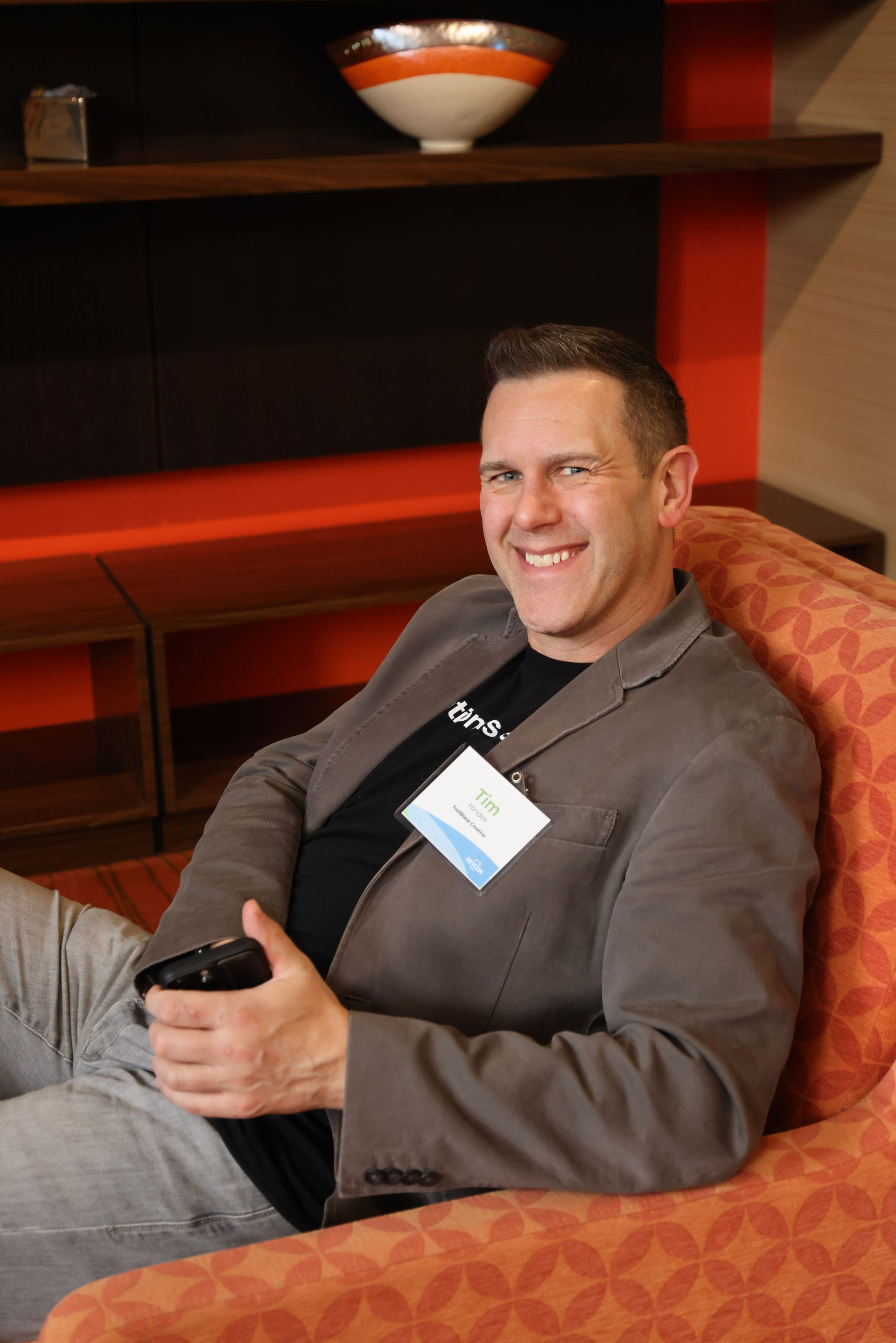 A smiling person wearing a blazer and name tag sits in an orange patterned chair in a room with orange decor.