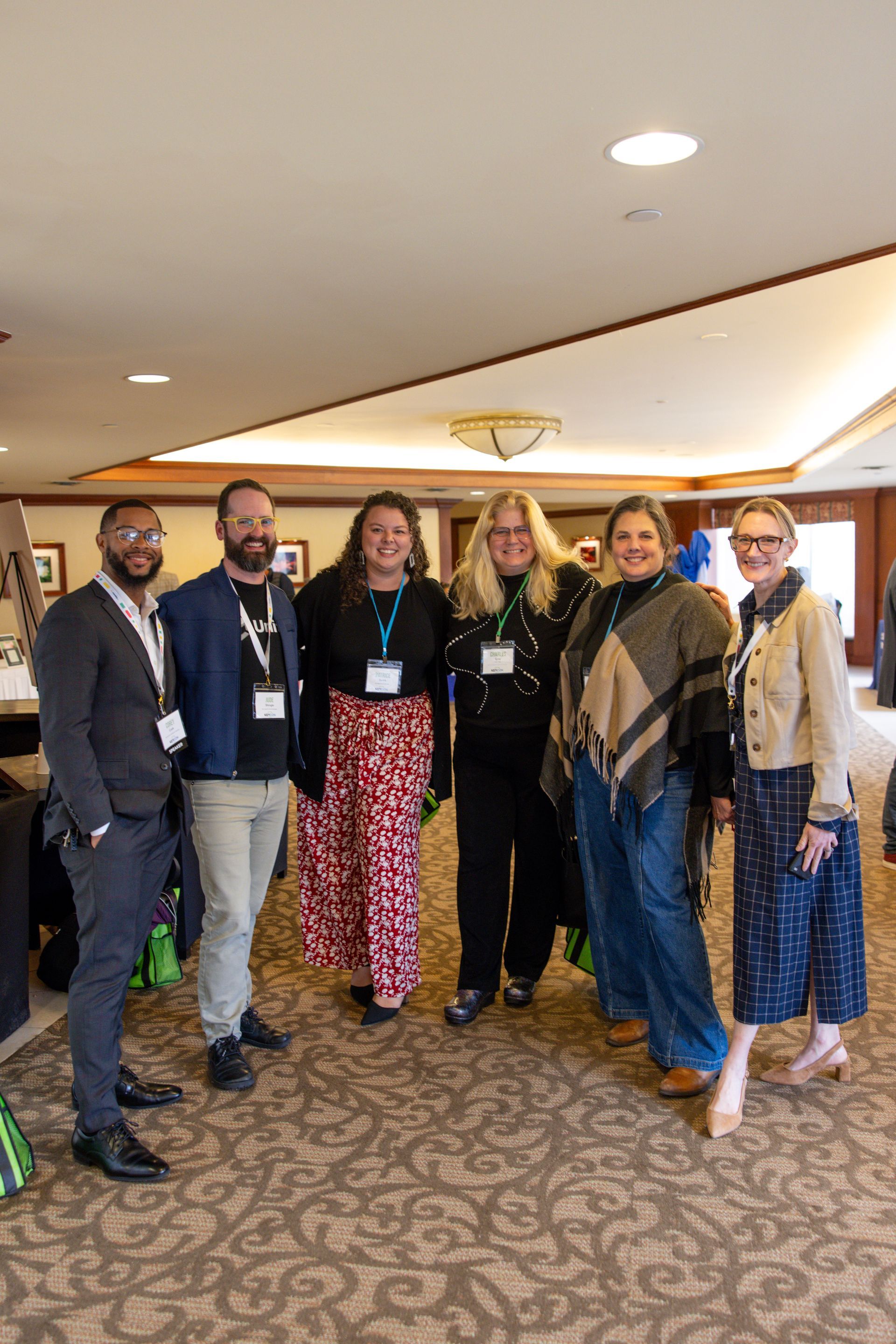 Six smiling individuals wearing event badges stand in a group for a photo in an indoor conference room setting.
