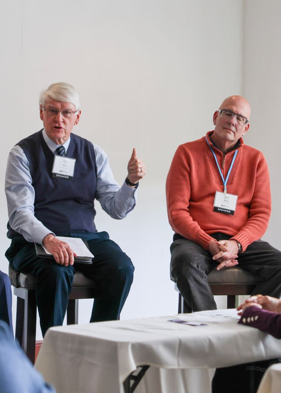 Two men in business attire sit at a table during a meeting; one is speaking with a hand gesture while the other listens.