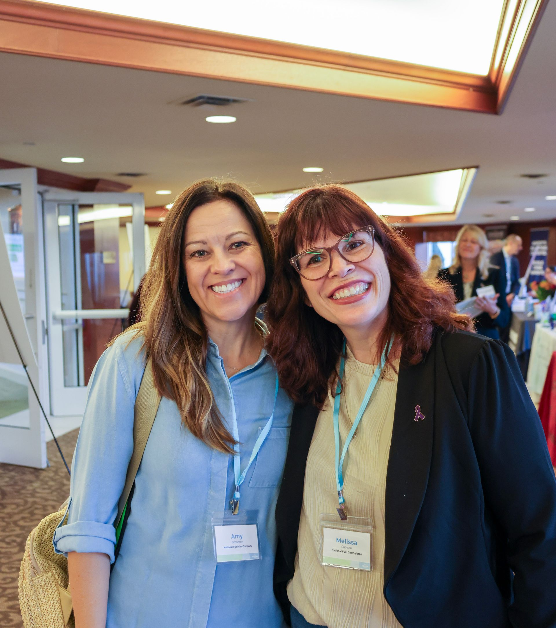 Two smiling women wearing name badges stand side-by-side in an indoor event space.