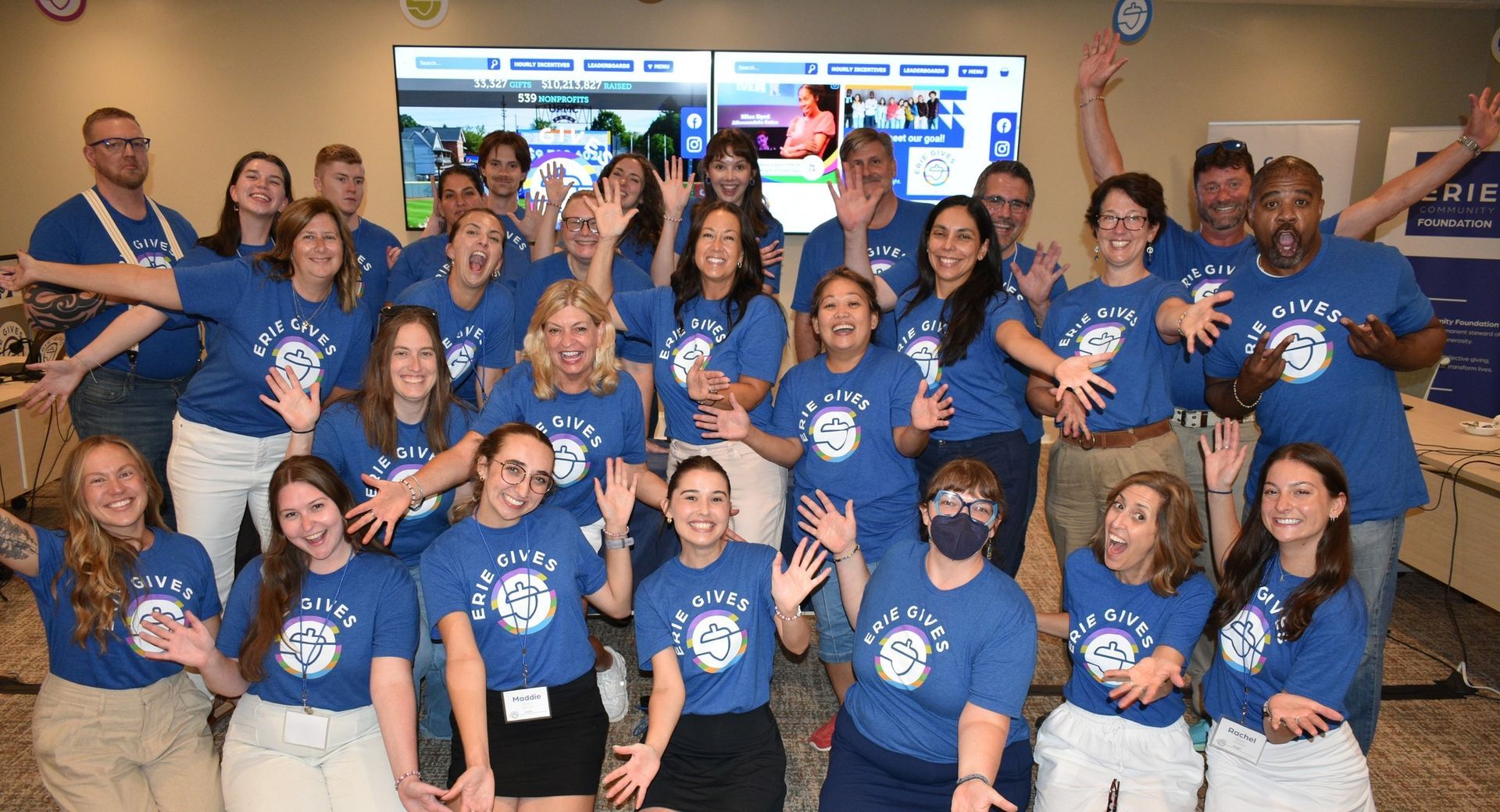 A large group of people in matching blue shirts pose together, smiling and waving in an indoor office setting.