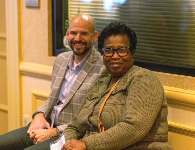 Two people sit side-by-side against a window, one in a plaid blazer and the other in a textured olive jacket.