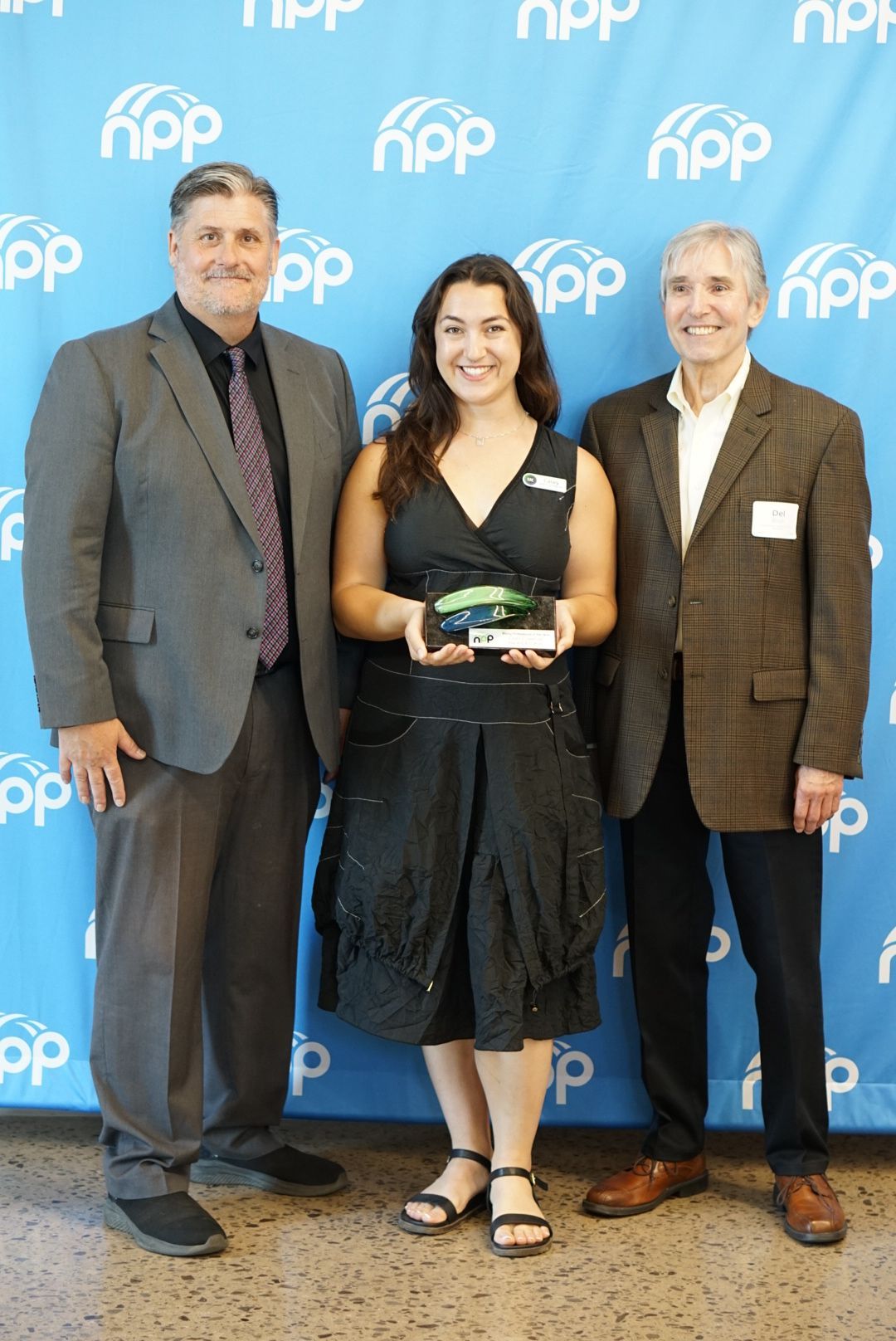 A woman holding an award stands between two people in suits in front of a blue background with the white 