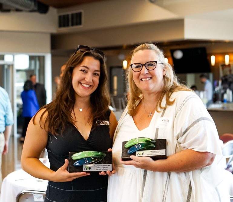 Two smiling individuals stand side-by-side in a room, each holding an award plaque with green and blue glass accents.