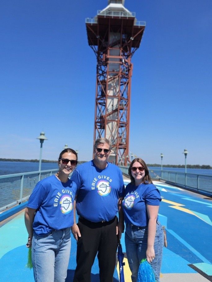 Three people in matching blue shirts pose on a blue-painted pier with the Bicentennial Tower in Erie, PA, behind them.