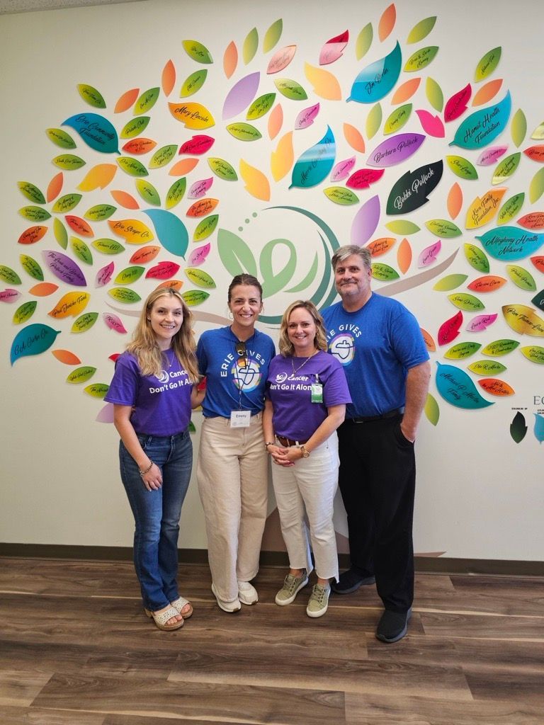 Four people in blue and purple shirts stand smiling in front of a colorful wall mural of a tree with leaf-shaped plaques.