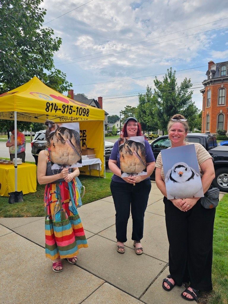 Three people stand on a sidewalk near a yellow tent, holding large photos of birds in front of their faces.