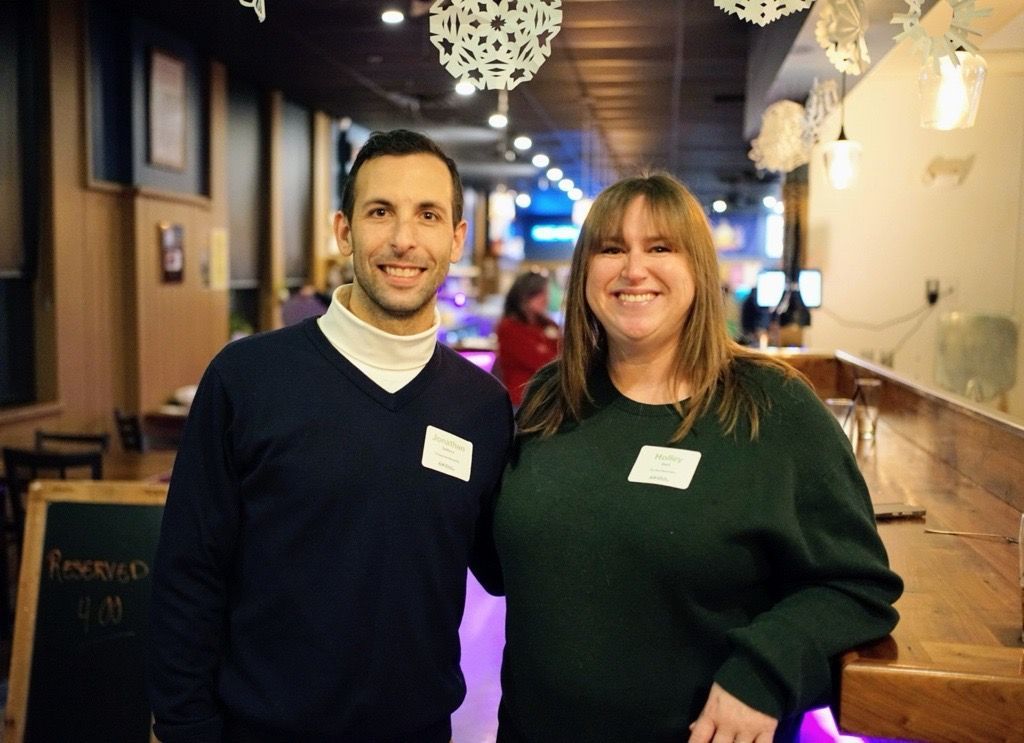 A man and woman wearing name tags and casual sweaters pose for a photo indoors, standing near a wooden counter.