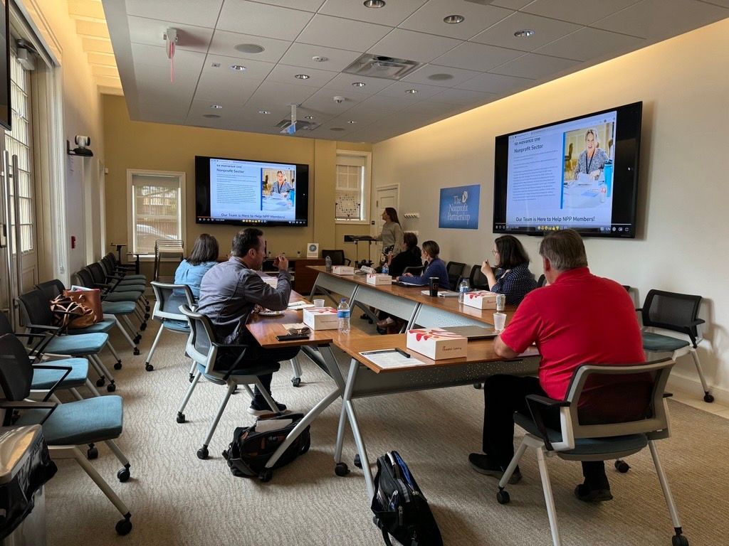 A small group sits around a conference table in a bright room during a meeting, with two screens displaying presentation.