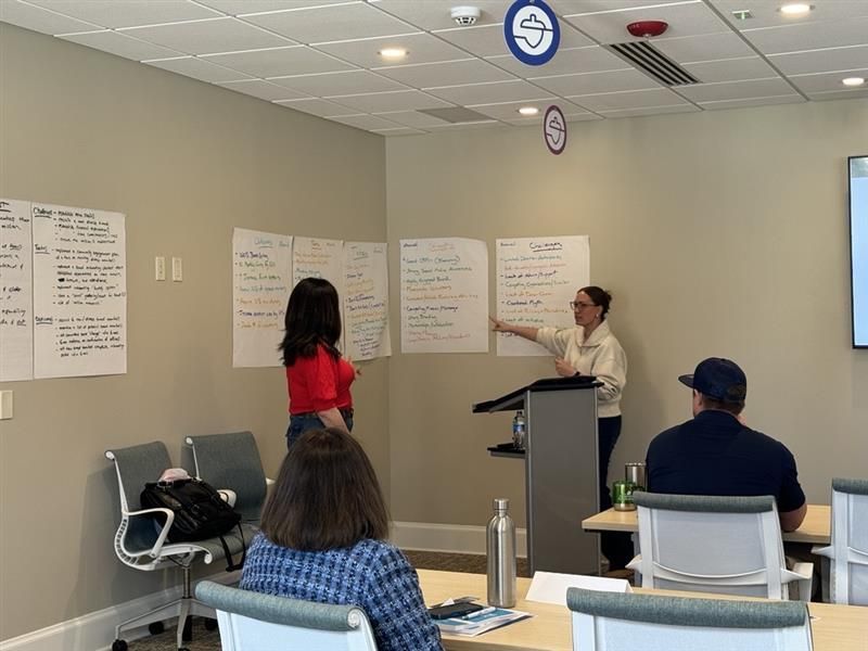Two people stand before wall-mounted posters presenting to a group in a brightly lit conference room.