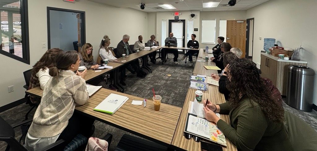 A group of people sit around a U-shaped table in a bright conference room for a meeting, with laptops and papers.