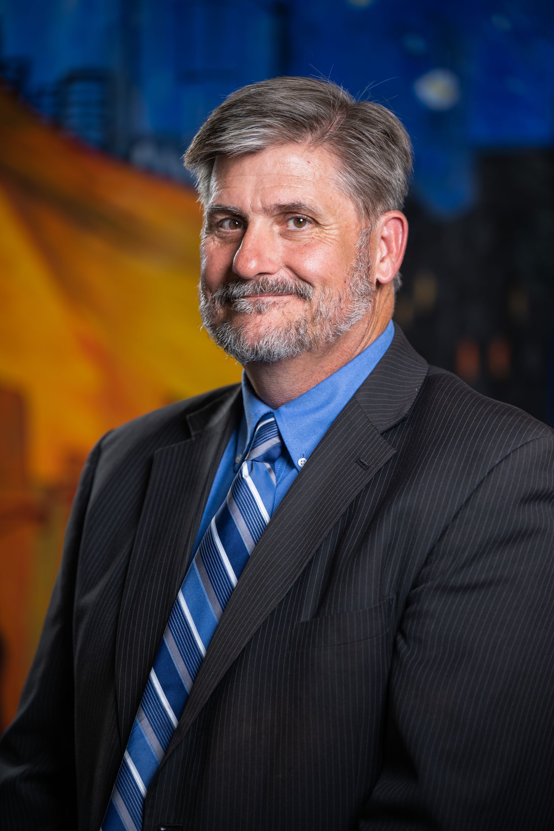 A professional portrait of a man wearing a dark textured suit, blue shirt, and blue striped tie against a blurry background.