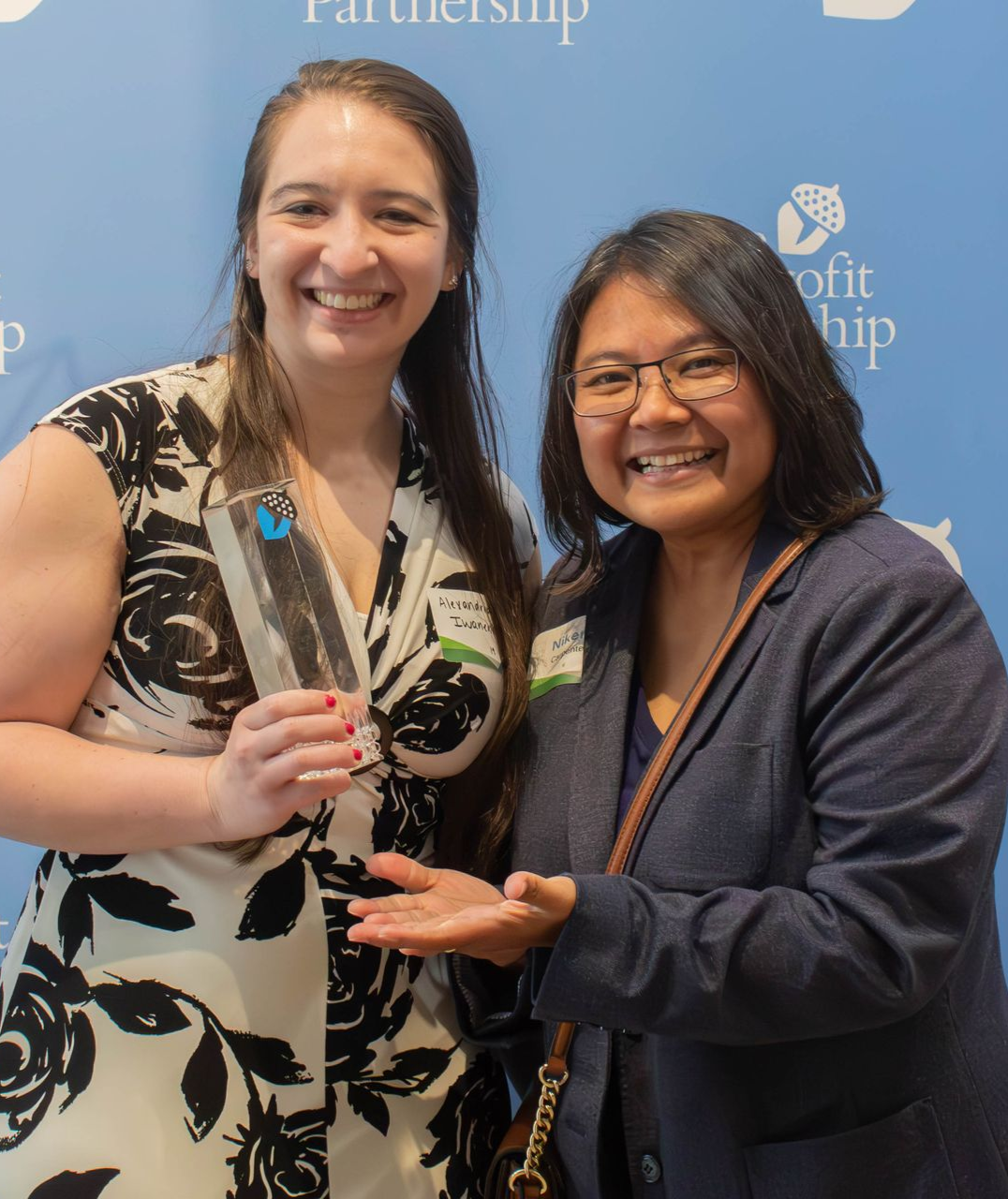 Two smiling people pose for a photo in front of a branded blue backdrop, one holding an award and the other gesturing to it.