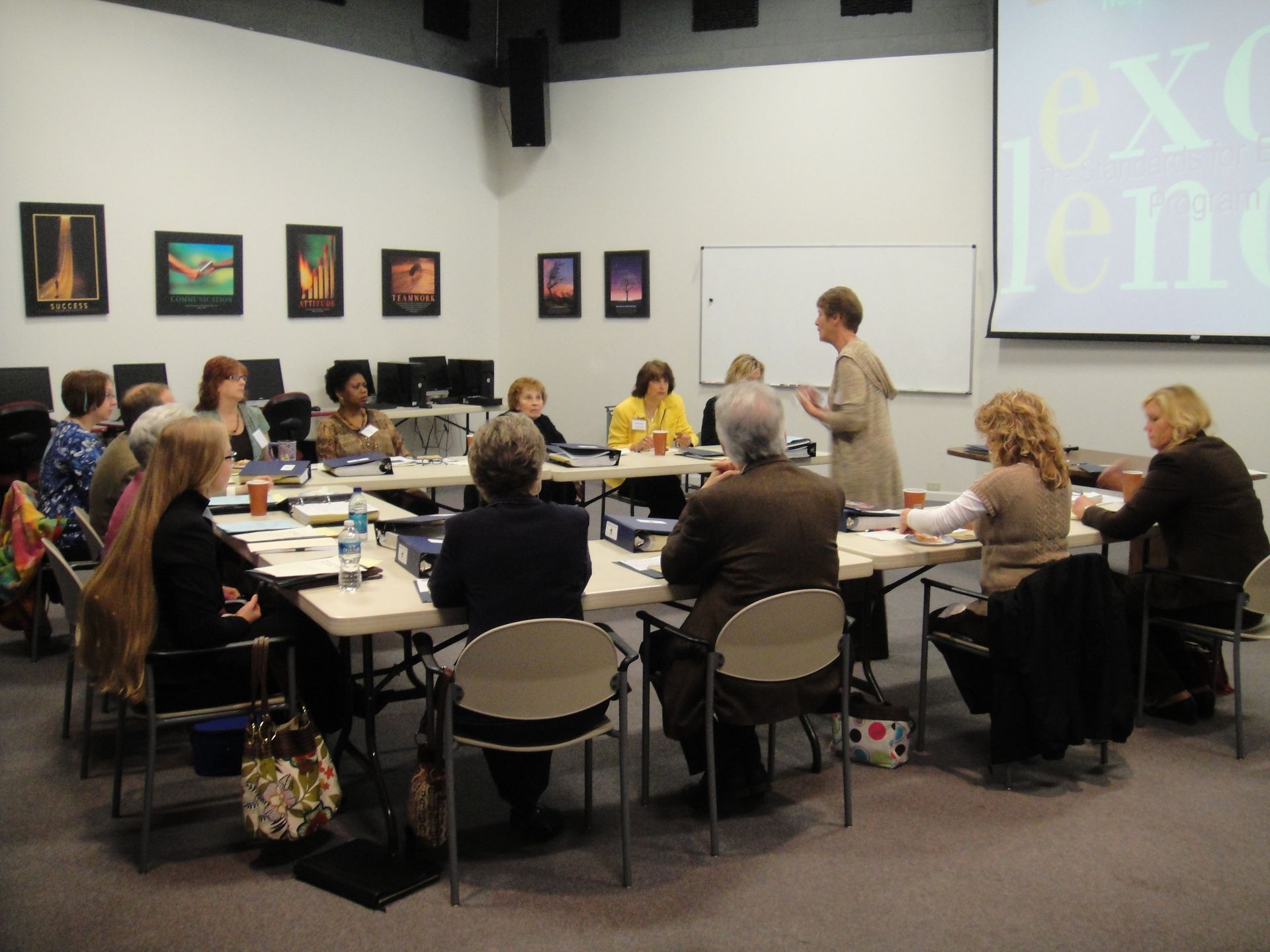 A presenter leads a meeting for a group of people seated around U-shaped tables in a bright, modern conference room.