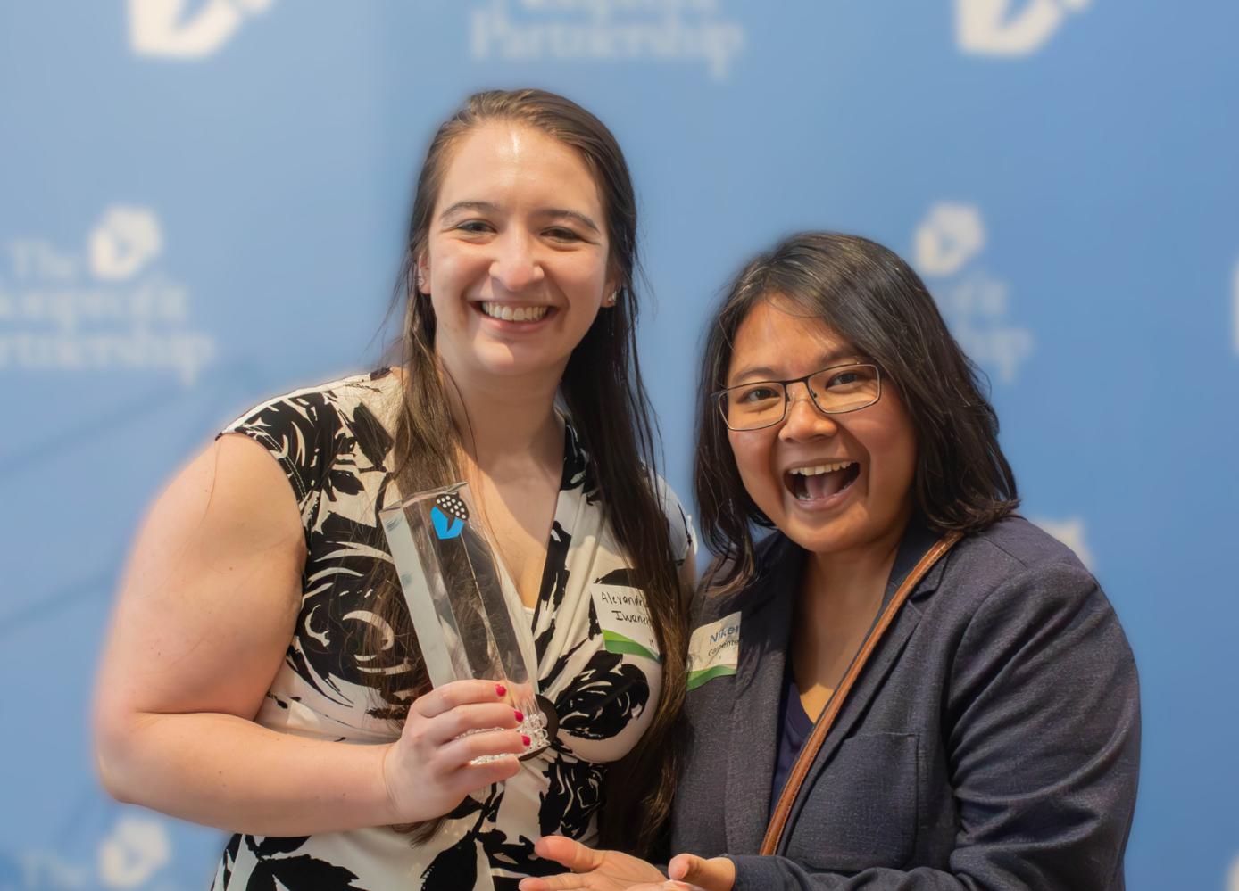 Two smiling people stand in front of a blue banner, with one holding a glass trophy.