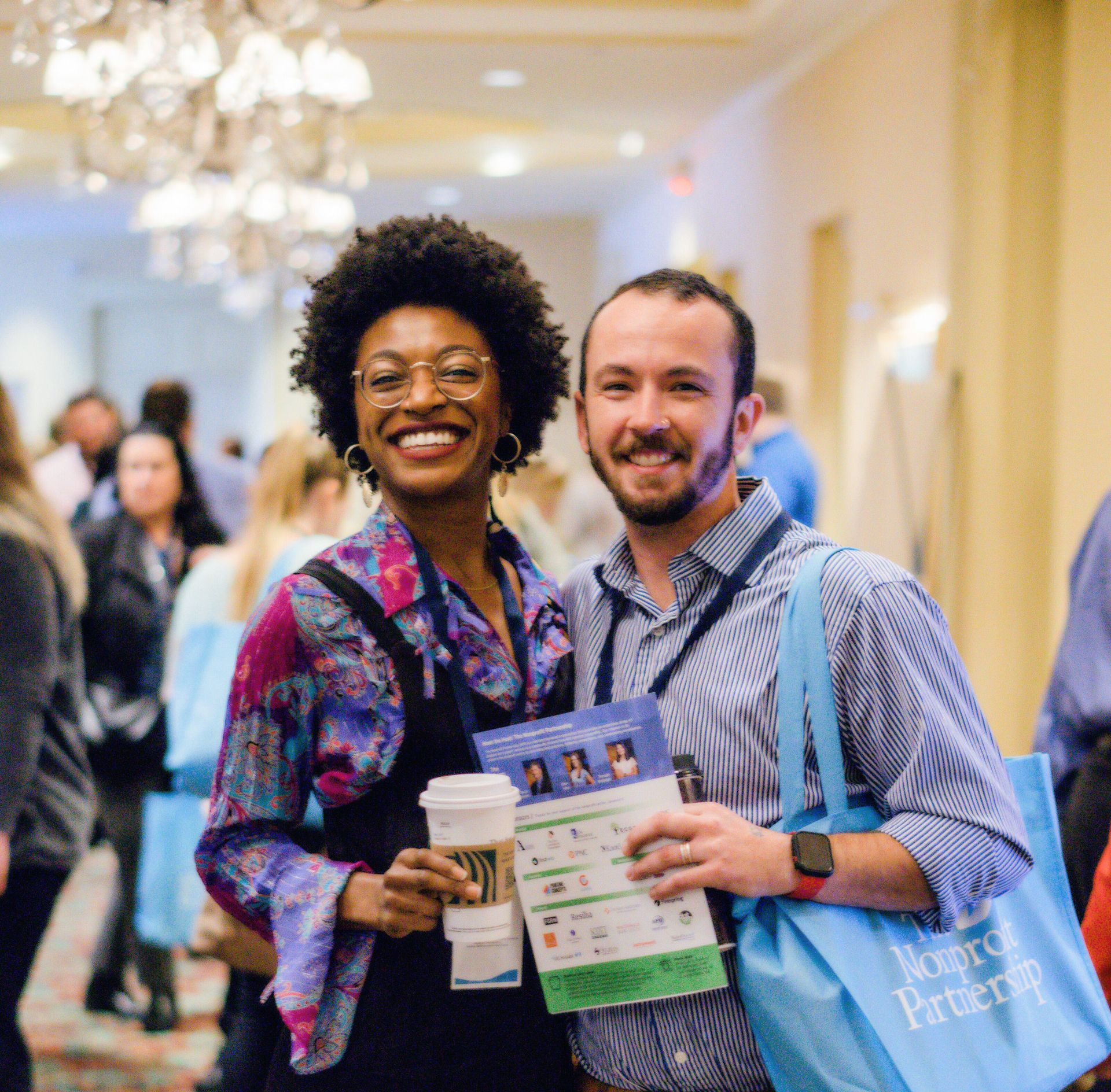 Two smiling professionals pose in a conference hall, holding coffee and event materials while carrying branded tote bags.