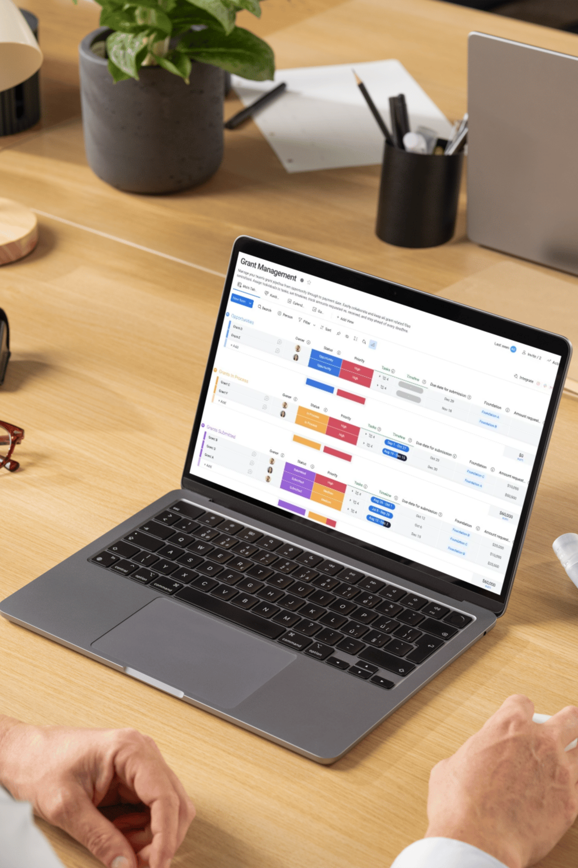 Hands typing on a laptop displaying a project management dashboard on a wooden desk with a plant and office supplies.