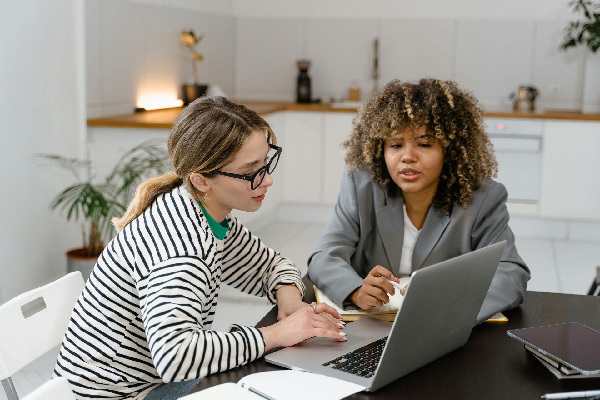 Two people collaborating on a laptop at a kitchen table. One types while the other observes and speaks.